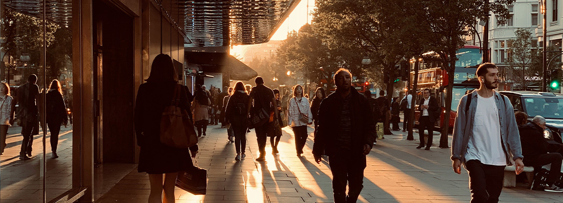 City street scene at sunset with people walking on a sidewalk lined with trees and buildings. The setting sun casts long shadows, and a double-decker bus is visible in the background.